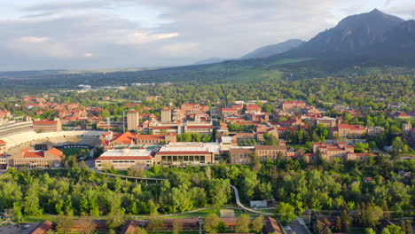 aerial pan up reveal of beautiful flatiron mountain vista, bright green trees, and cu boulder campus in boulder colorado during an evening sunset with warm light on the summer landscape