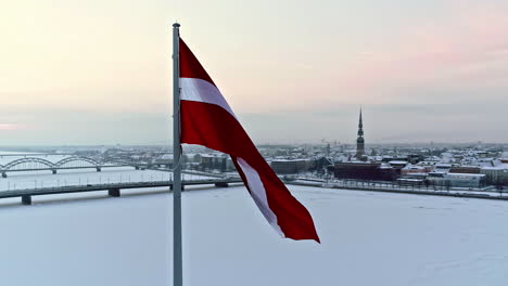 orbiting aerial view of a latvian flag waving in the wind over a snowy, winter city and frozen river