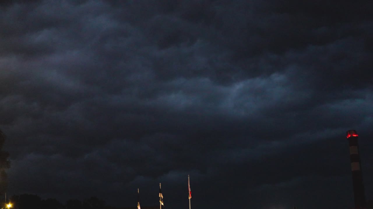 Dramatic bottom up view of dark blue and black thunder stormy clouds moving above an urban area during night time while lighting strikes in the background, large smoke chimney stack in the foreground