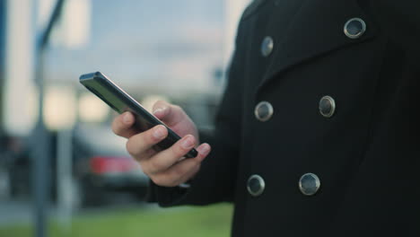 close-up of individual in black coat multitasking while holding coffee cup and operating phone outdoors, showcasing modern lifestyle and focus in urban setting with blurred greenery