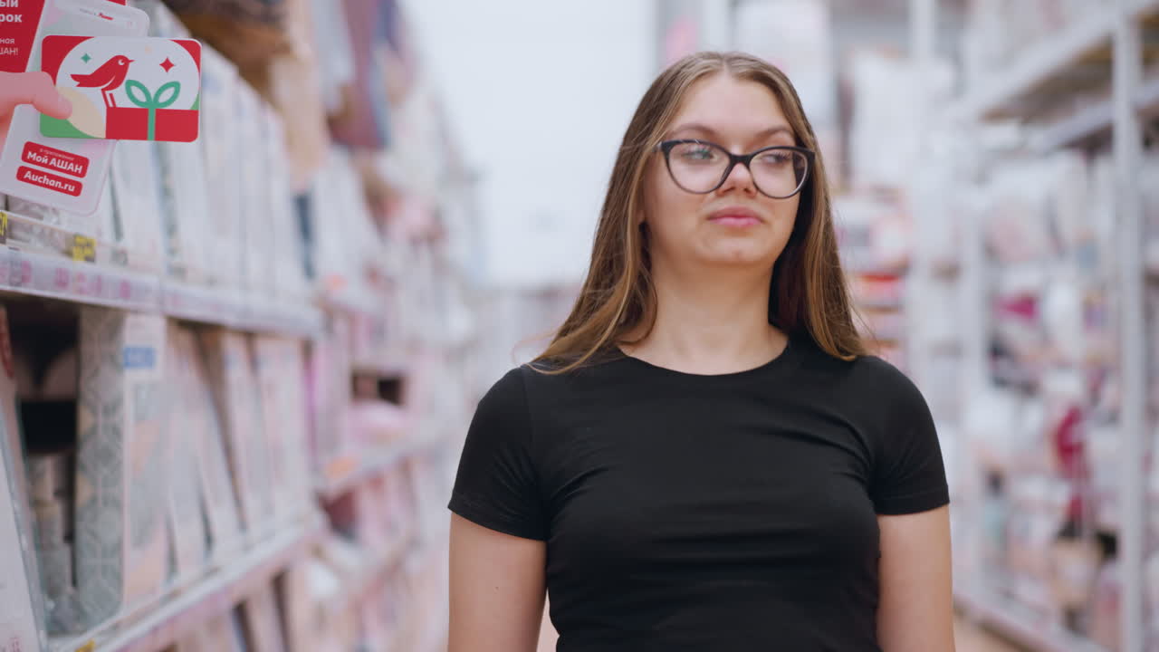 estudiante con camiseta negra y gafas caminando por el pasillo del supermercado mirando las mercancías en el lado izquierdo antes de dirigir la atención a la derecha, estantes y pantallas abastecidas con varios productos en el fondo