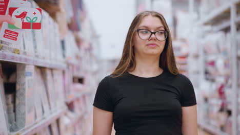 estudiante con camiseta negra y gafas caminando por el pasillo del supermercado mirando las mercancías en el lado izquierdo antes de dirigir la atención a la derecha, estantes y pantallas abastecidas con varios productos en el fondo