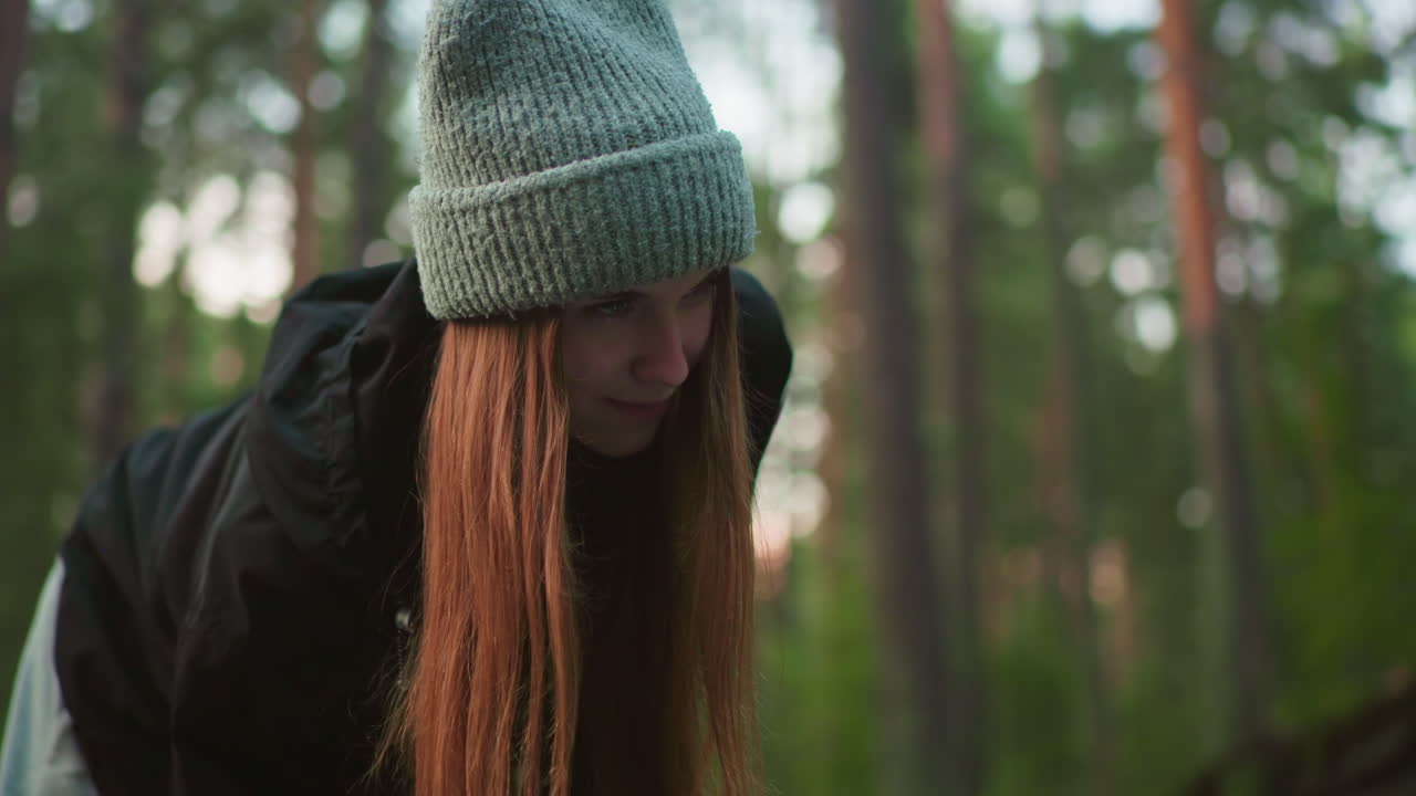 young woman wearing knitted cap and black jacket glances up with cheerful expression while focusing on outdoor task in forest with blurred trees and soft natural lighting