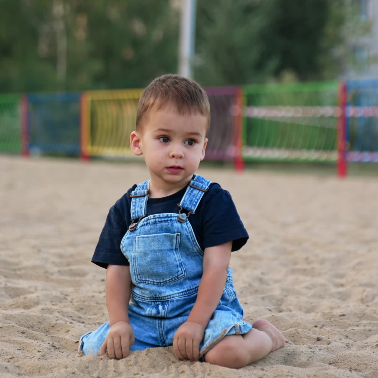 Cute baby boy in jeans romper sits in the sand outdoors. Kid deeps his hands into sand looking attentively away