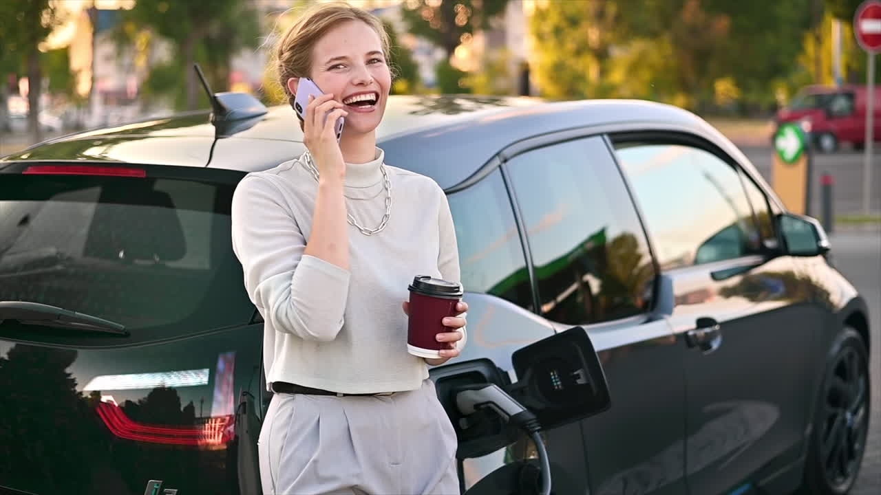 Una giovane donna bionda con un caffè parla al telefono a una stazione di ricarica per auto elettriche con un'auto elettrica nelle vicinanze a Chișinău al tramonto, Moldavia. Rallentatore