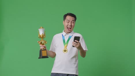 asian man with a gold medal and trophy looking at smartphone then screaming goal celebrating on green screen background in the studio