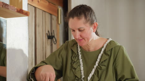 focused cook wearing green dress and white apron stirs pot on stovetop while wooden kitchen door and wall mirror reflect her movements, capturing calm home cooking atmosphere