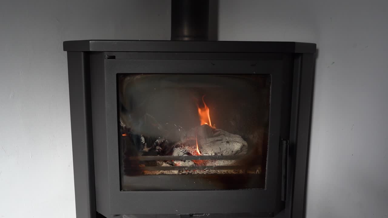 A fire burns brightly in a black stove. The flames dance as they consume logs placed inside. Smoke rises through the chimney. The room is quiet and minimal