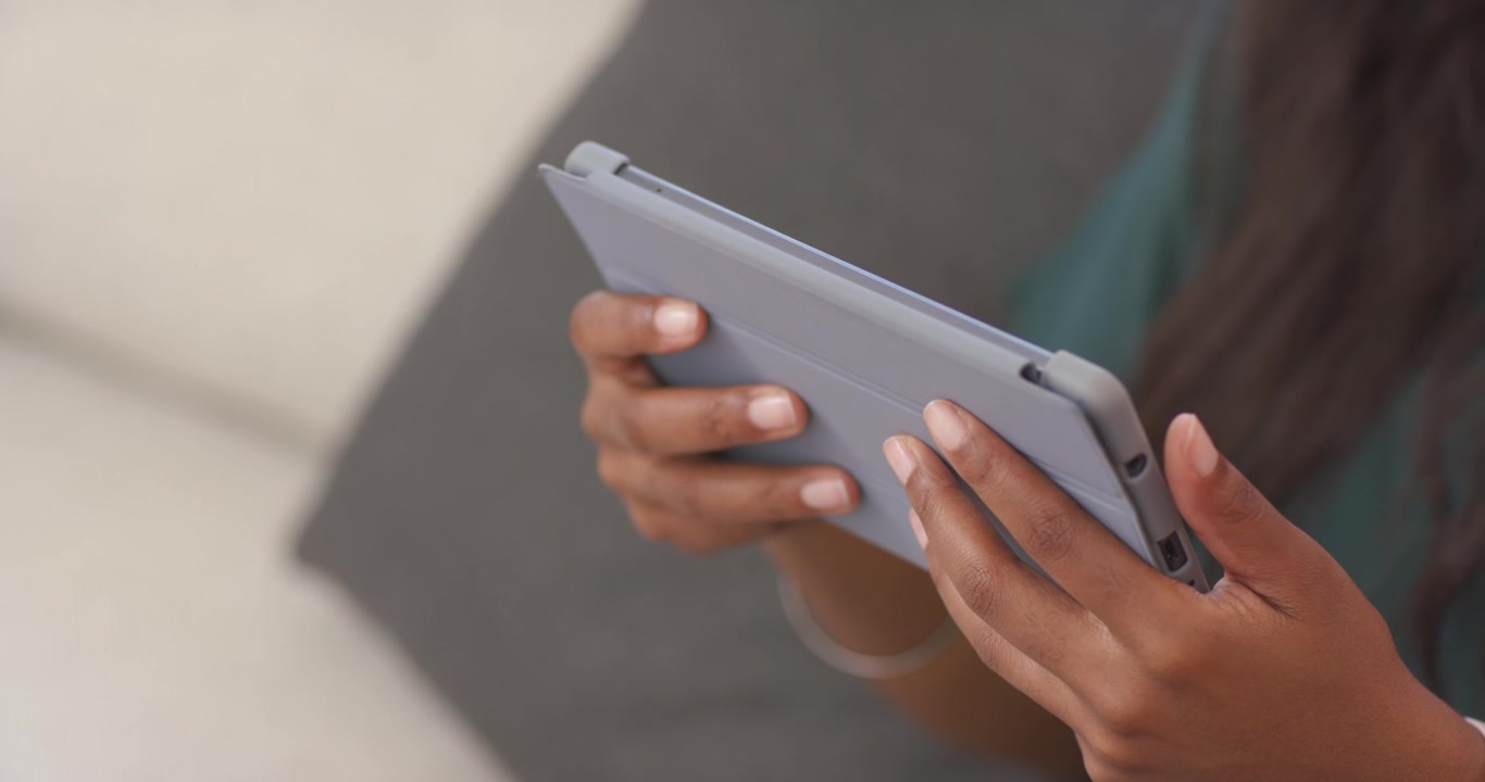 Mulher africana adulta segurando uma capa tipo fólio de tablet, tocando com o polegar e ajustando a pegada em um sofá azul-petróleo.