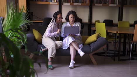 Two-stylish,-focused-female-friends-sitting-on-grey-and-yellow-couch-with-one-laptop,-studying-or-browsing-smth-together.-Two-students-working-remotly-with-one-laptop.-Modern-interior