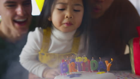 Cerca-De-Una-Familia-Con-Dos-Padres-Celebrando-El-Cumpleaños-De-Su-Hija-En-Casa-Apagando-Las-Velas-Del-Pastel