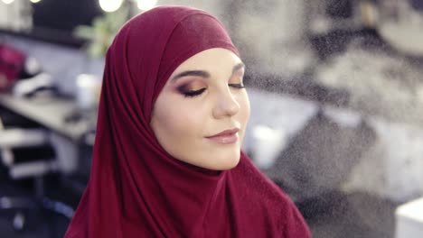 hermosa joven con ojos de avellana y hijab púrpura en la cabeza tiene maquillaje de fijación de niebla rociado en su cara mientras obtiene los toques finales de maquillaje impecable