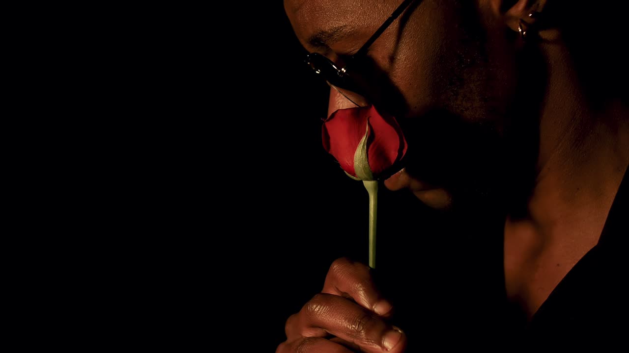 Stylish man smelling - rubbing rose against cheeks, profile close up, black background
