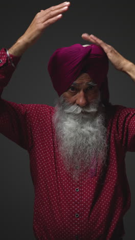 vertical video low key studio lighting shot of senior sikh man with beard tying fabric for turban against dark background