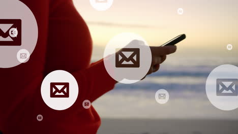 animación de iconos de correo sobre la sección media de una mujer biracial que usa un teléfono inteligente en la playa
