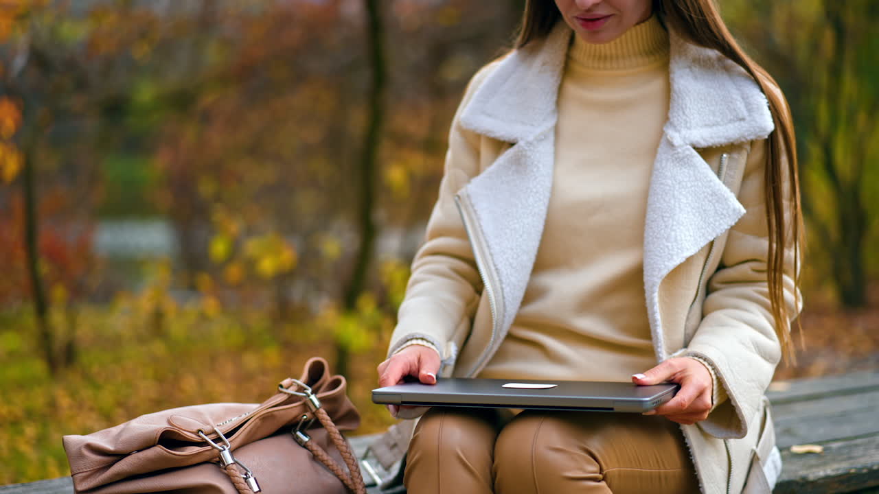 Attractive woman sitting outdoors on the wooden table opens her laptop. Freelancing lady starting work in the nature in autumn park.