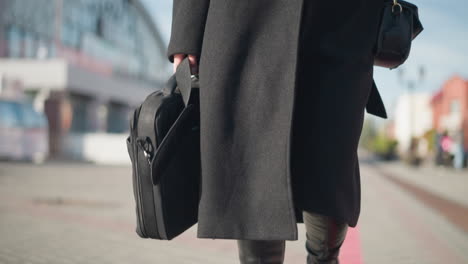 close-up of black bag held by person wearing stylish black coat and leather boots, walking on sunny urban street with blurred background details of modern architecture and pedestrians in motion