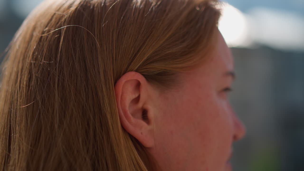 Close up head view of woman with golden hair adjusting hair outdoors under warm sunlight, gentle motion of strands and soft focus background create serene mood