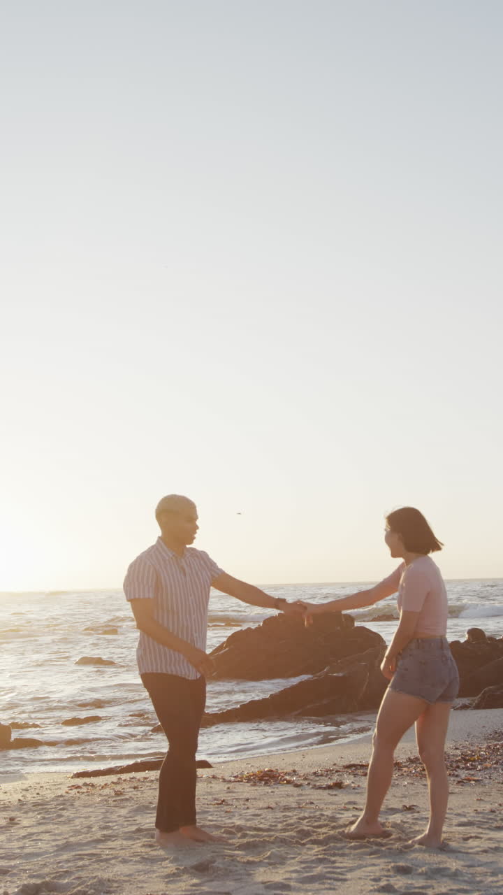 Vertical video of happy biracial couple dancing together at beach, in slow motion