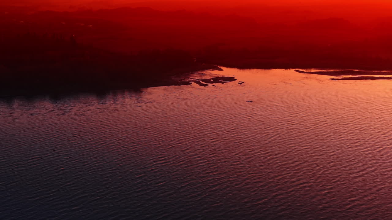 Red dusk reflections on lake surface. Close view of rippled water tinted crimson, dark treeline in distance
