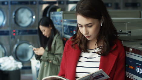 caucasian young pretty and stylish girl sitting in laundry service room. beautiful woman reading magazine and flipping pages while waiting for clothes to be washed.