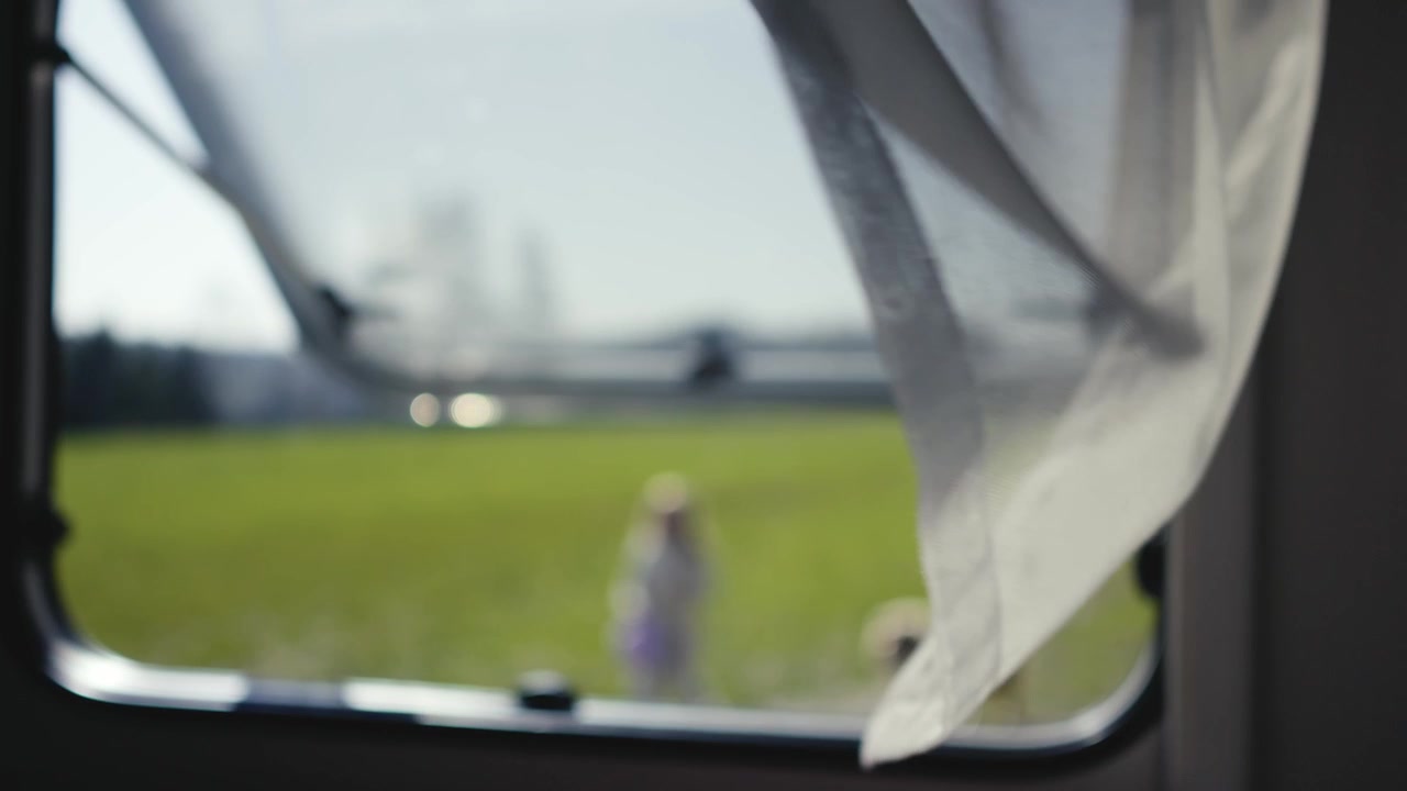 Slow motion shot of a billowing curtain at the open window of a caravan, with a blurred view of children playing in a meadow in the background