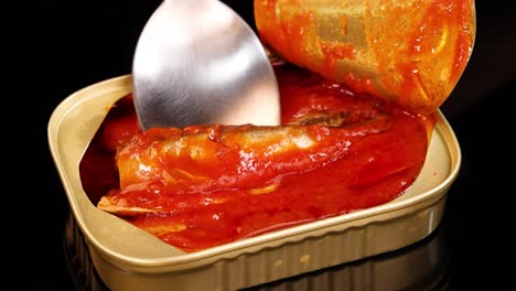 a metal spoon lifts a sardine from an open tin filled with tomato sauce under bright, even lighting. close-up, static camera, food-focused composition