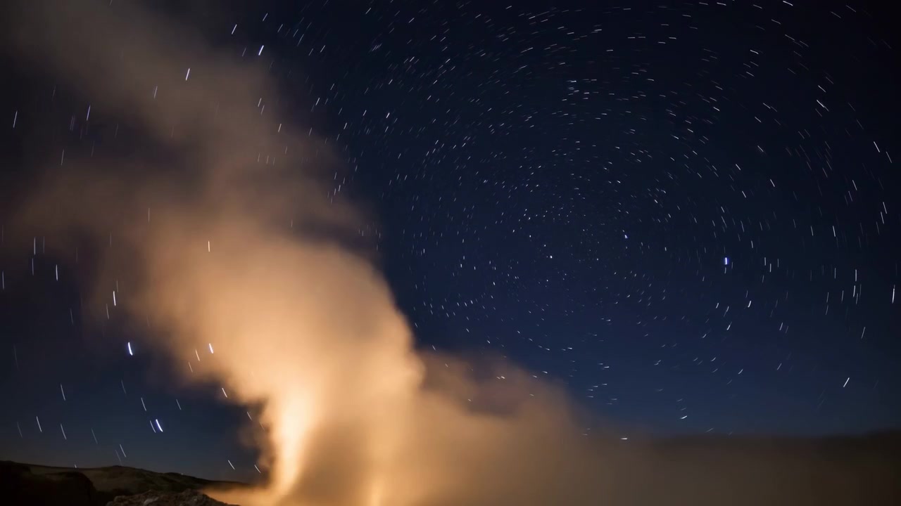 Luminous steam plume rises from lower left and spreads into deep night sky while circular star trails sweep across upper right above low dark landscape silhouette, with night, sky, star, trail detail