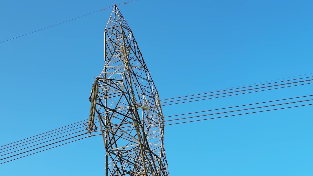 Close view of steel transmission tower with insulators and high voltage power lines cutting across clear blue sky, emphasizing grid infrastructure and industrial design