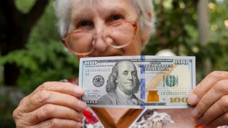 old woman in eyeglasses showing one hundred dollar bill into camera and smiling outdoor. granny holding cash outside. money concept. close up slow motion