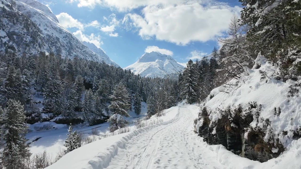 Camminata in prima persona lungo un sentiero in una valle innevata con un fiume ghiacciato e pini in una soleggiata giornata invernale.