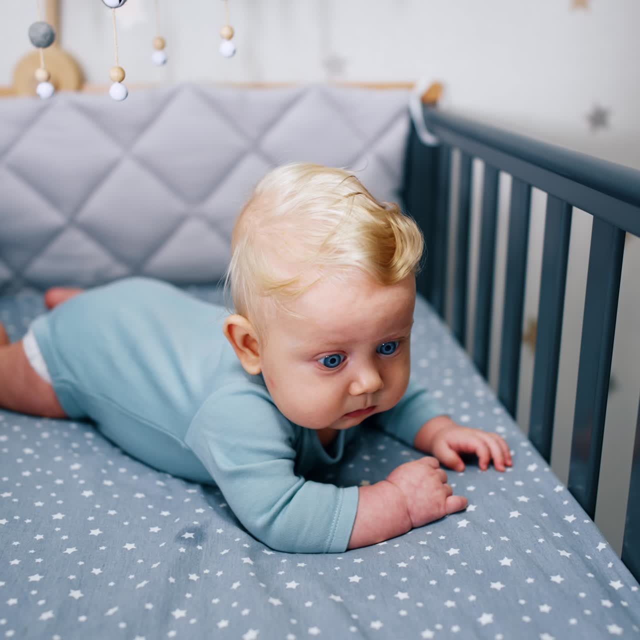 Adorable blond baby boy in blue bodysuit lying in the crib. Calm blue-eyed child looking intently ahead