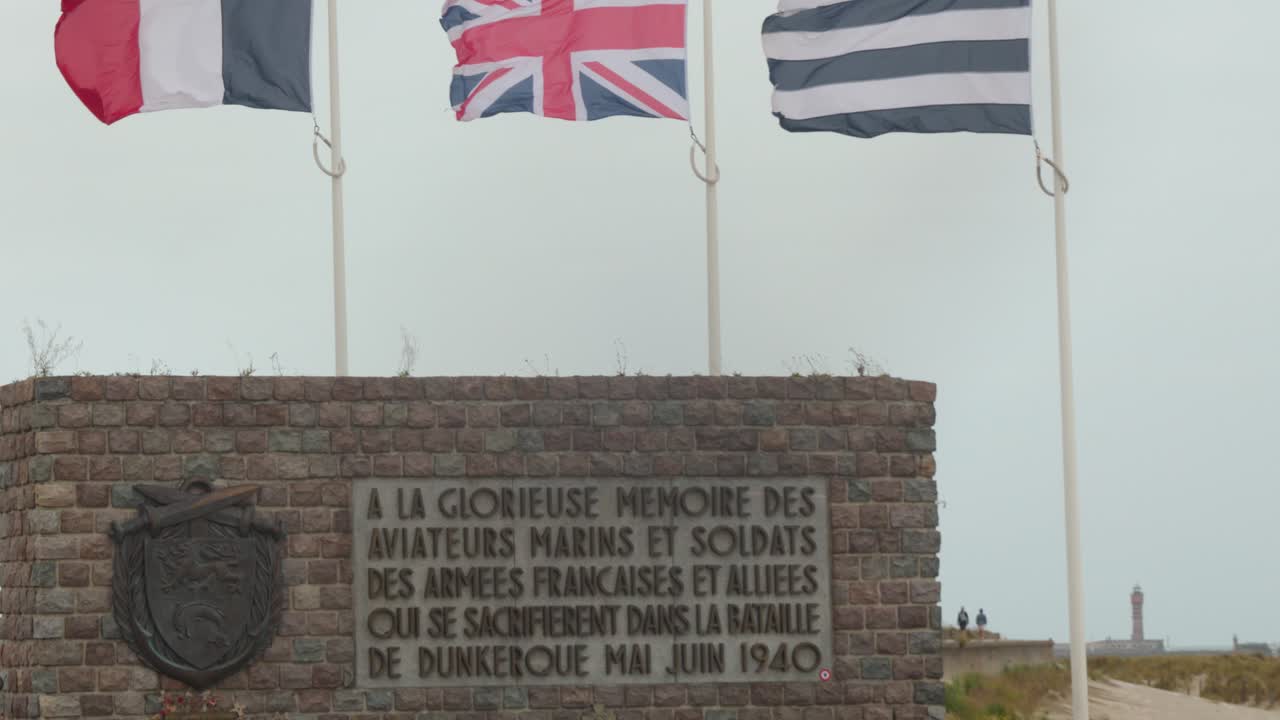 Camera pans right across Dunkirk WWII memorial, brick monument, and fluttering Allied flags, overcast daylight