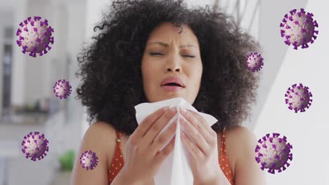 animation of coronavirus cells rotating over biracial woman with afro hair sneezing with tissue