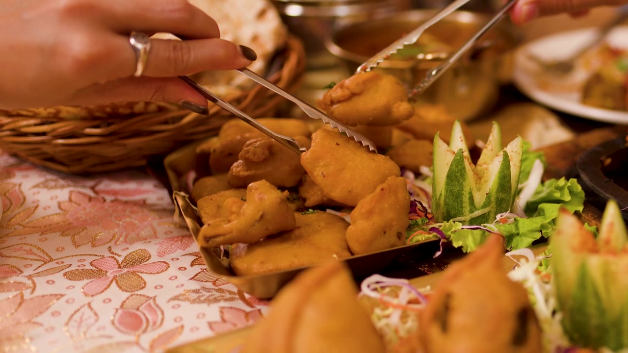 Primo piano di mani che servono pakora e samosa con le pinze durante un pasto tradizionale indiano.