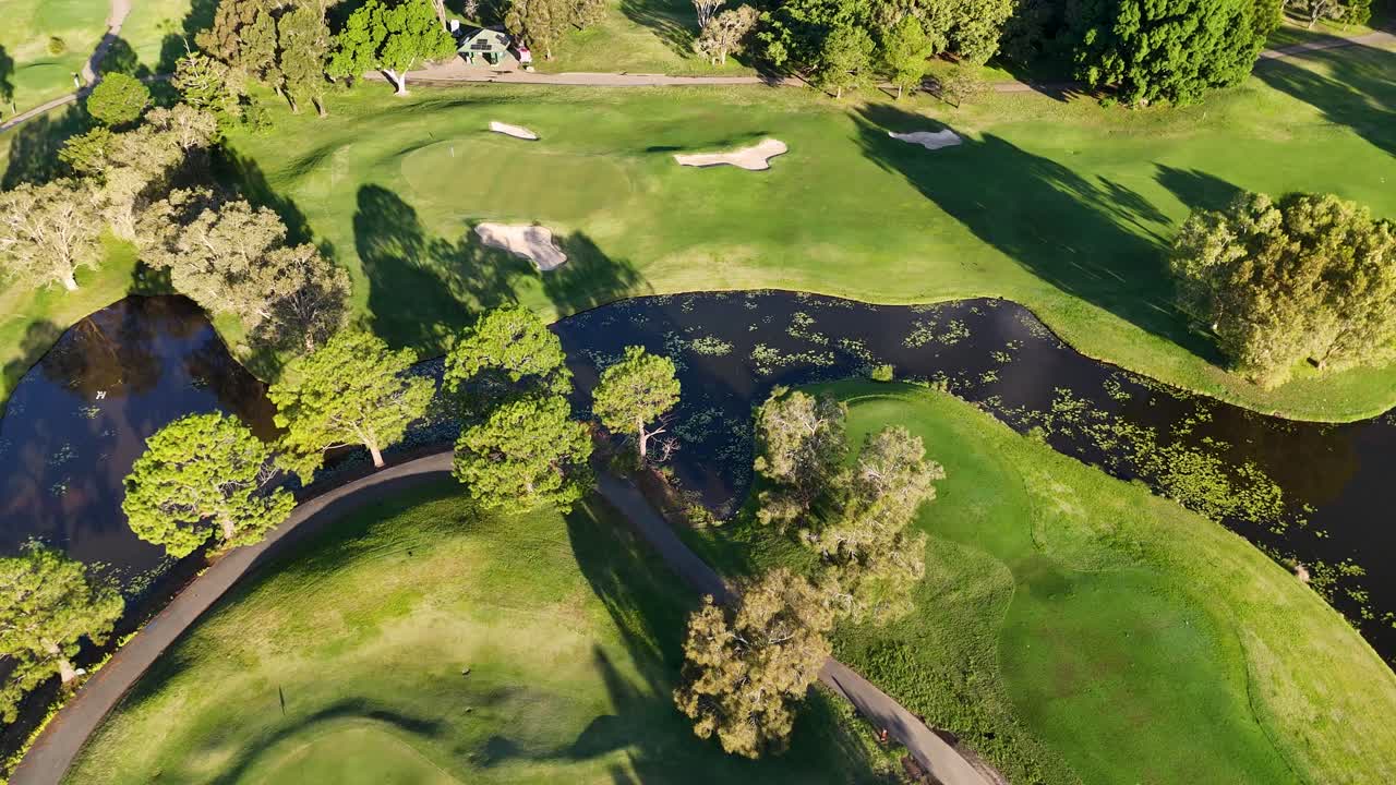 Drone footage captures a lush golf course with fairways, bunkers, and water hazards under bright sunlight