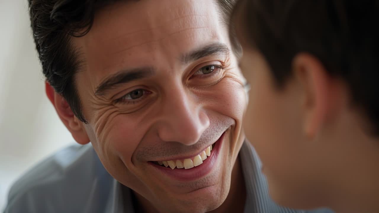 Leaning father in light-blue collared shirt smiling and meeting child's gaze at home, showing love