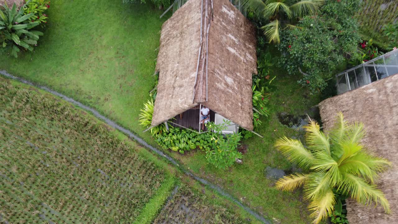 una persona sentada en la terraza de una cabaña de paja con techo de paja en medio de un entorno natural rural, bali