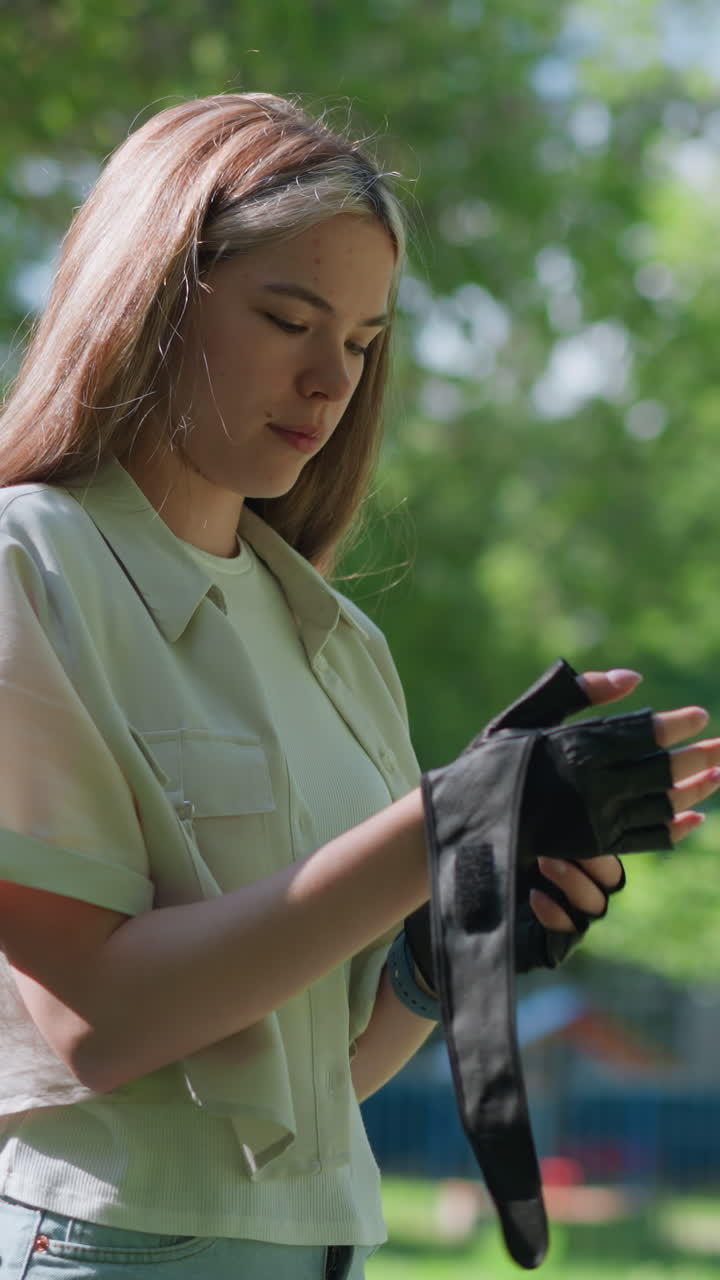 Young woman standing outdoors, thoughtfully removing biker glove from right hand, with lush greenery and trees in blurred background, background includes rail and park elements