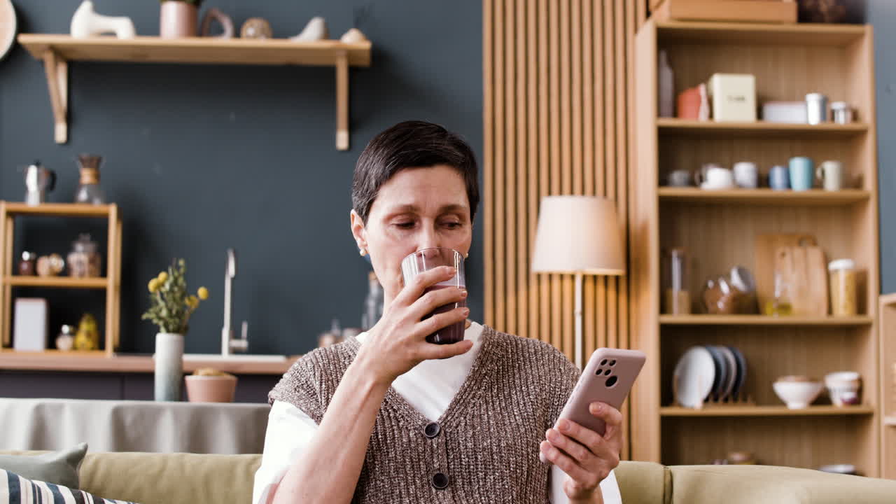Kvinne slapper av hjemme, drikker en drikke og bruker smarttelefonen sin i sofaen.
