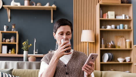 woman relaxing at home, drinking a beverage and using her smartphone on the couch.