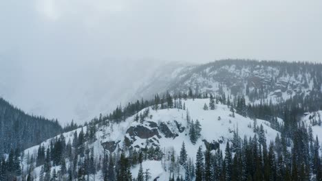 an aerial drone shot moving backwards while slowly panning sideways overlooking foggy mountains with trees covered in snow during the winter