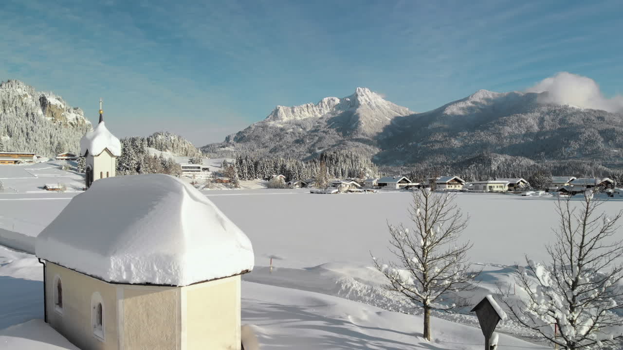 Aerial Drone shot showing a snowy landscape in in the sunshine of the Tyrol, Austria and revealing a snow covered tiny chapel in Lechaschau