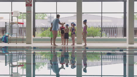 Entrenador-Masculino-Dando-Instrucciones-A-Los-Niños-En-La-Clase-De-Natación-Mientras-Están-Parados-En-El-Borde-De-La-Piscina-Cubierta