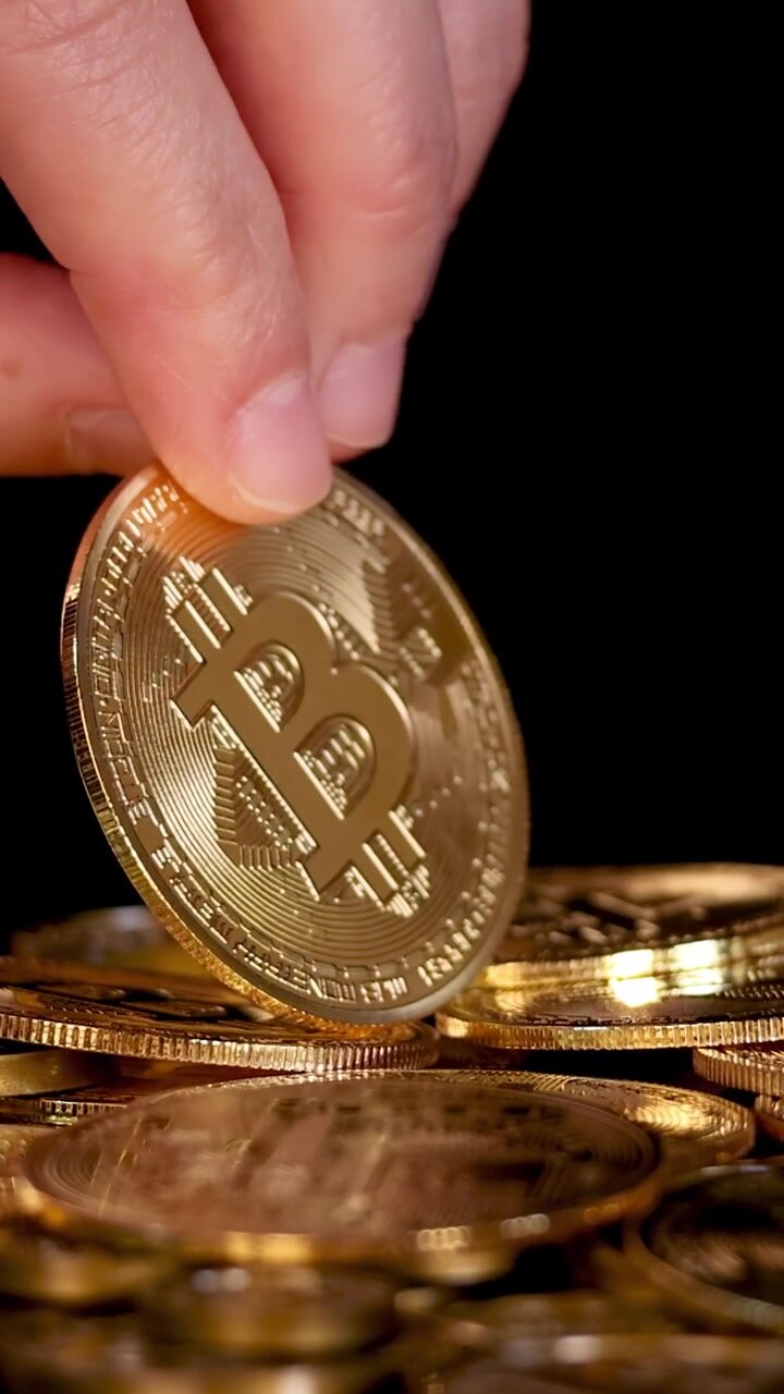 Una mano lascia cadere un token Bitcoin d'oro su una pila di monete, primo piano, illuminazione drammatica, sfondo nero.