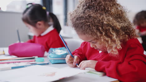 close up of mixed race schoolgirl drawing at desk in an infant school classroom, focus on foreground