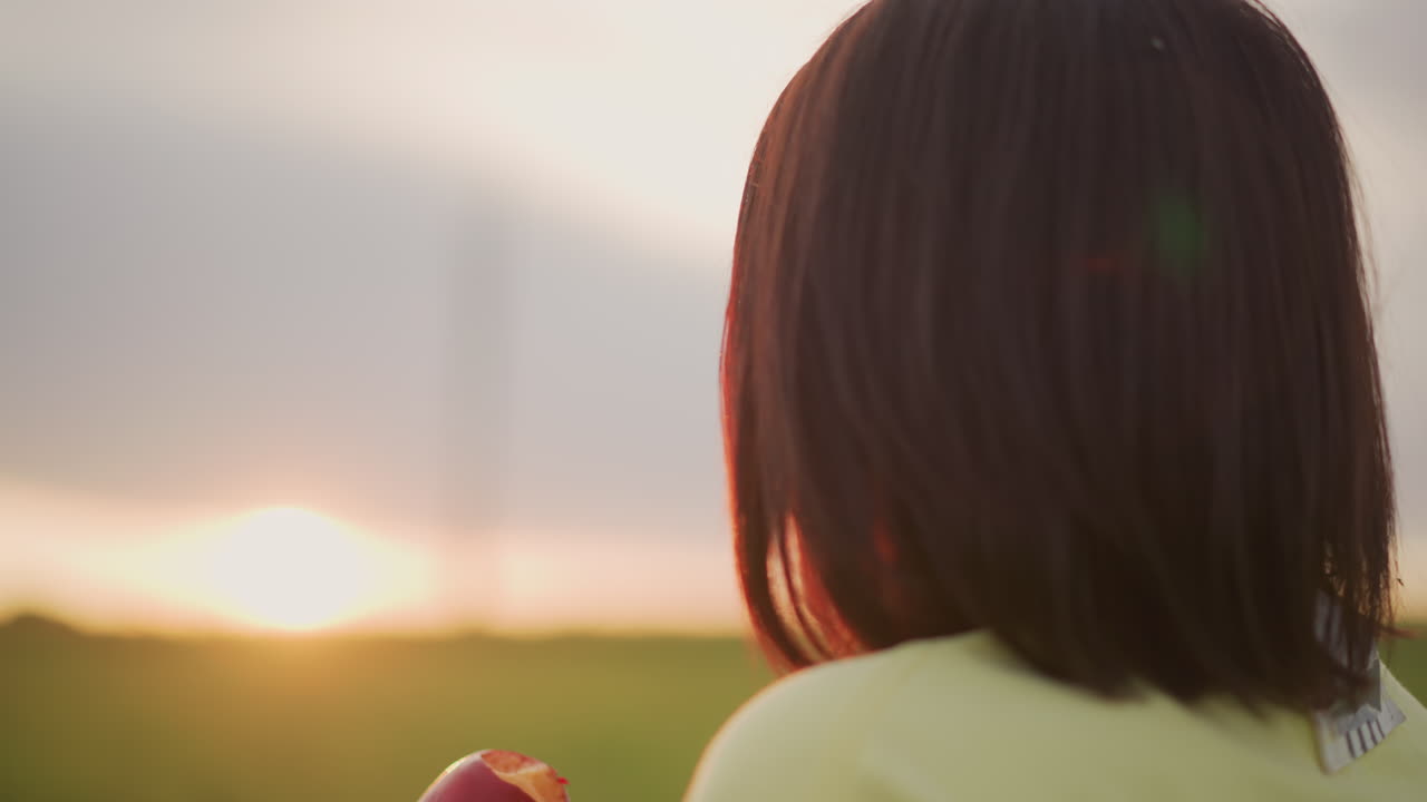 해 질 녘에 사과를 천천히 베어 물고 있는 백인 여성의 옆모습, 황금빛 시간, 부드러운 역광 실루엣, 초원 위에 은은한 플레어, 사색에 잠긴 표정, 공부를 멈춘 학생, 생각에 잠긴 작가