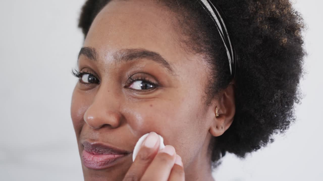 Close up of happy african american woman cleansing face with cotton pad in bathroom, slow motion
