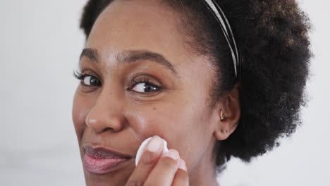 primer plano de una feliz mujer afroamericana limpiando la cara con una almohadilla de algodón en el baño, cámara lenta