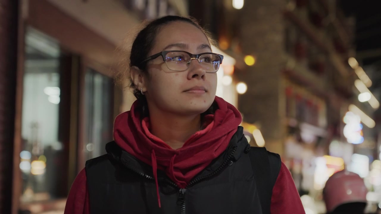 Donna asiatica che cammina di notte per una strada del centro illuminata al neon, indossando una felpa rossa con cappuccio e occhiali, espressione contemplativa, profondità di campo ridotta con luci bokeh calde, atmosfera urbana tranquilla